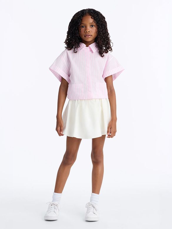 Young girl models a cute pink striped short-sleeve shirt with a white pleated skirt. Perfect kids' fashion for summer outfits.