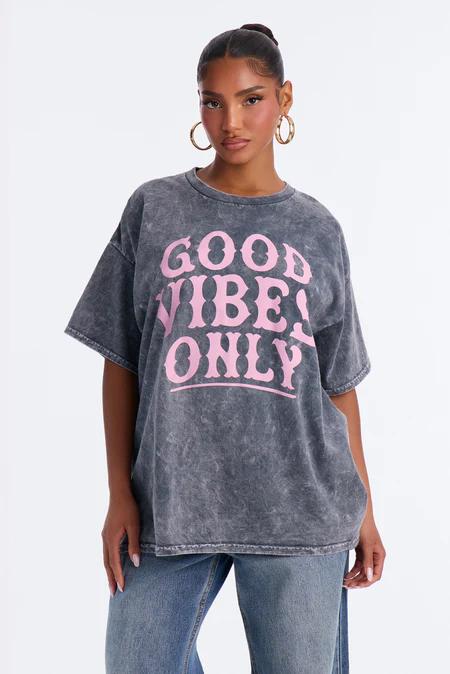 Woman in an oversized charcoal acid-wash t-shirt with prominent pink "GOOD VIBES ONLY" text, light blue jeans, and gold hoop earrings.