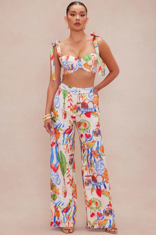 Woman models a vibrant fruit print two-piece set: bustier crop top with tied straps and flowy wide-leg pants. Perfect resort wear collection.
