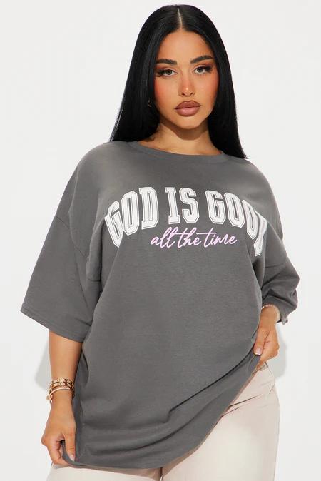 Mujer con camiseta holgada gris carbón con "GOD IS GOOD" en blanco y "all the time" en cursiva rosa. Lleva pulseras doradas.