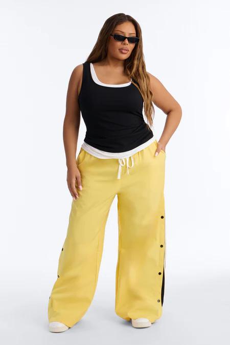 Gafas de sol negras, camiseta negra con ribete blanco, pantalones anchos amarillos con broches laterales y raya negra. Zapatillas blancas.
