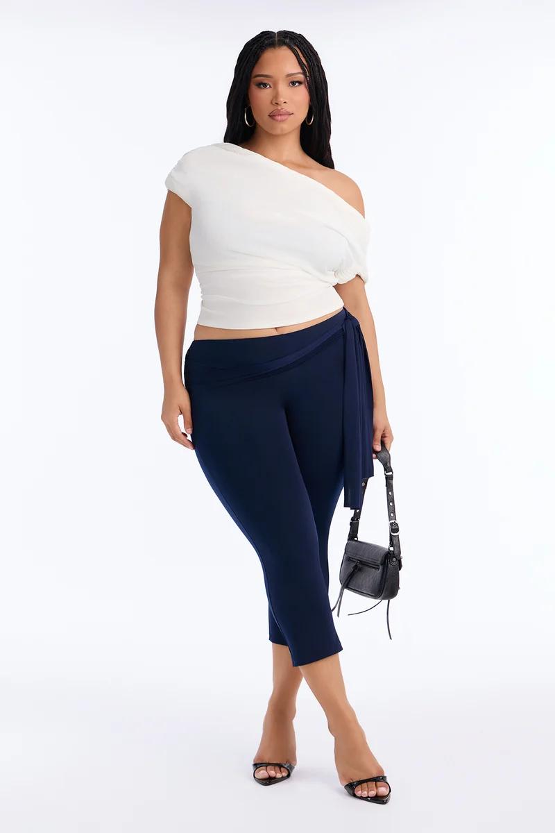 Plus size model wearing a white one-shoulder draped top and navy blue cropped capri pants.