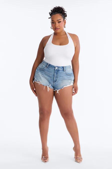 Woman with curly hair in a white halter top, light blue distressed denim shorts, and clear high-heeled mules on a white background.