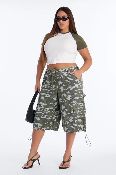 Mujer con top corto blanco y mangas verde oliva, shorts cargo camuflaje, sandalias negras de tacón y gafas de sol, sosteniendo un bolso negro.