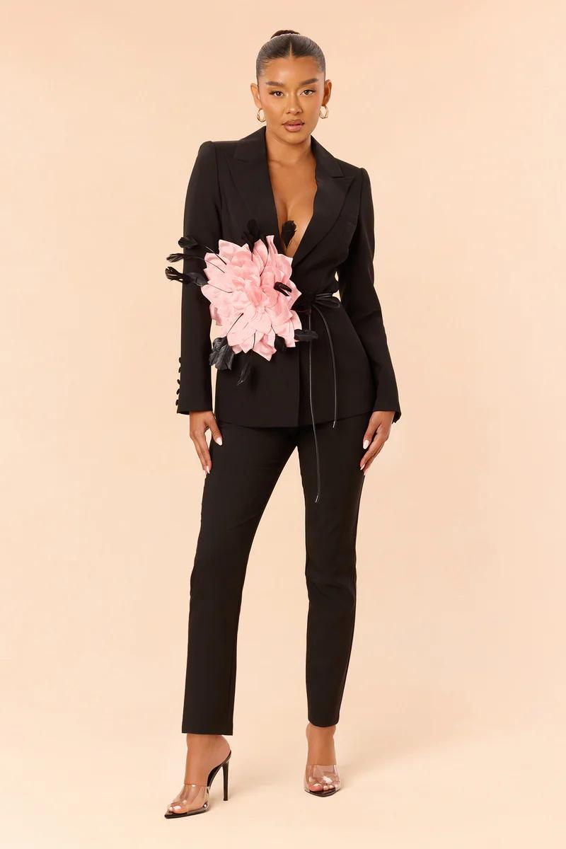 Woman in a chic black tailored suit with a large pink floral embellishment and black tie belt. Elegant women's fashion statement.