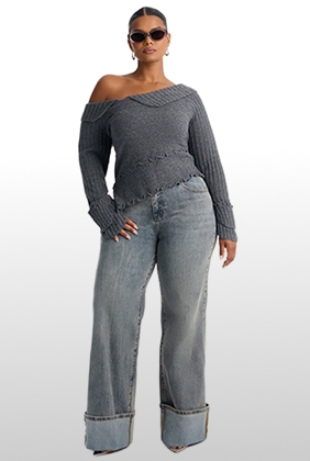 Modelo de talla grande con suéter gris de hombro caído y jeans anchos con vuelta, luciendo una nueva colección de moda femenina.