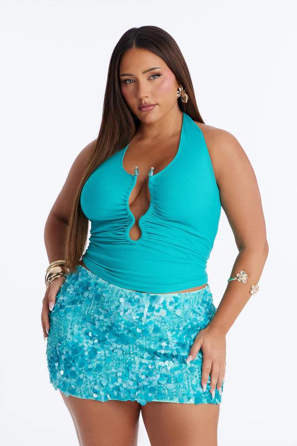 Woman in a teal halter top with a cutout and a matching sequin mini skirt. She wears gold bracelets, flower earrings, and an arm cuff.