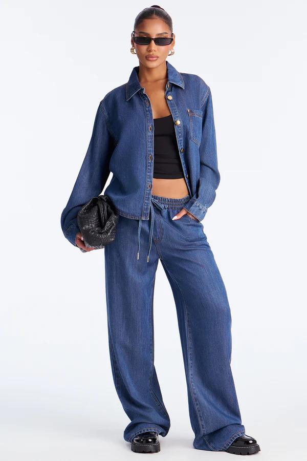 Modelo elegante con camisa vaquera azul holgada, top corto negro, pantalones de mezclilla de pernera ancha y bolso, presentando nueva colección casual.