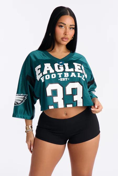 Woman in a cropped teal Eagles football jersey with white "EAGLES FOOTBALL EST 33" text, black shorts, and gold bracelets.