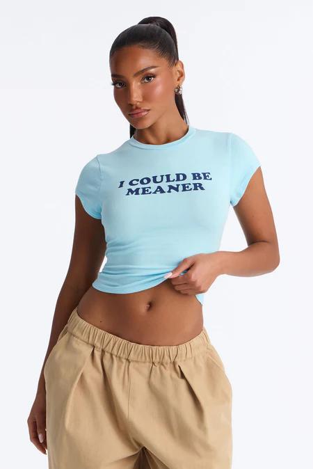 Model wearing a light blue crop top with "I COULD BE MEANER" text, paired with tan wide-leg pants. She pulls up the top showing her midriff.