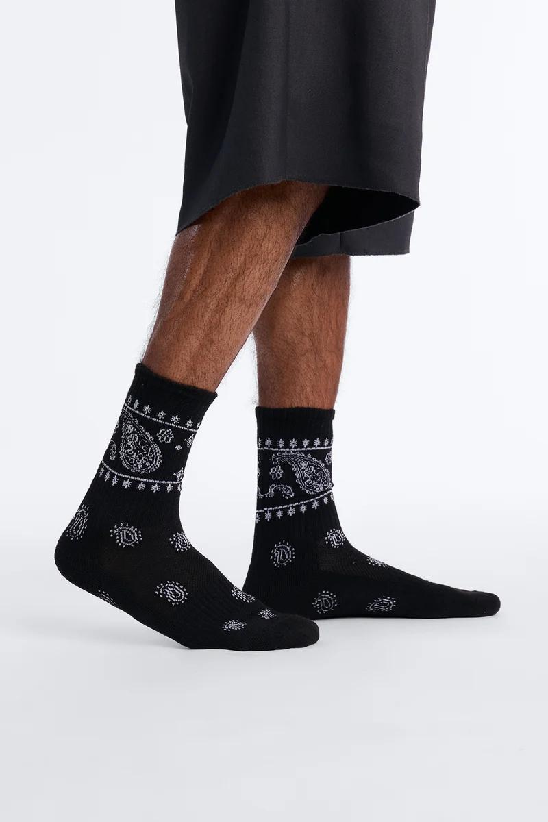 Lower legs and feet in black shorts and black crew socks with white paisley patterns. Shop our unique patterned socks collection for men.