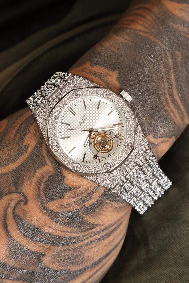 Reloj plateado brillante con diamantes, esfera blanca, engranajes visibles en brazo tatuado. Explora nuestra colección de relojes de lujo.