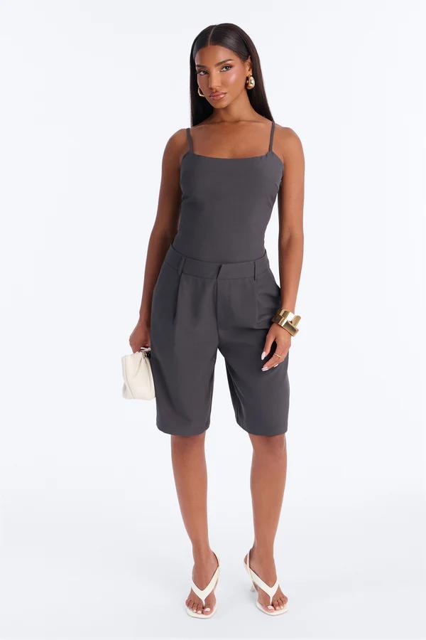 Woman in charcoal gray spaghetti strap top and matching pleated Bermuda shorts, holding a cream bag. Showcases a chic, versatile summer outfit.