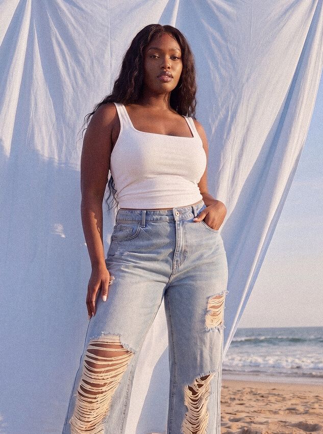 Woman modeling a white tank top and light wash ripped jeans against a beach backdrop with draped white fabric, highlighting casual summer style.