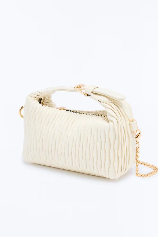 Elegante bolso de mano color crema fruncido con asa corta, cadena dorada visible y cremallera dorada. Inclinado sobre fondo blanco.