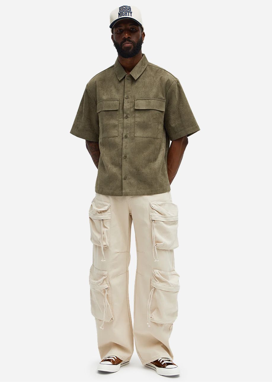 Hombre con camisa utilitaria verde oliva de manga corta, pantalones cargo color crema y gorra. Descubre la nueva colección streetwear para hombre.