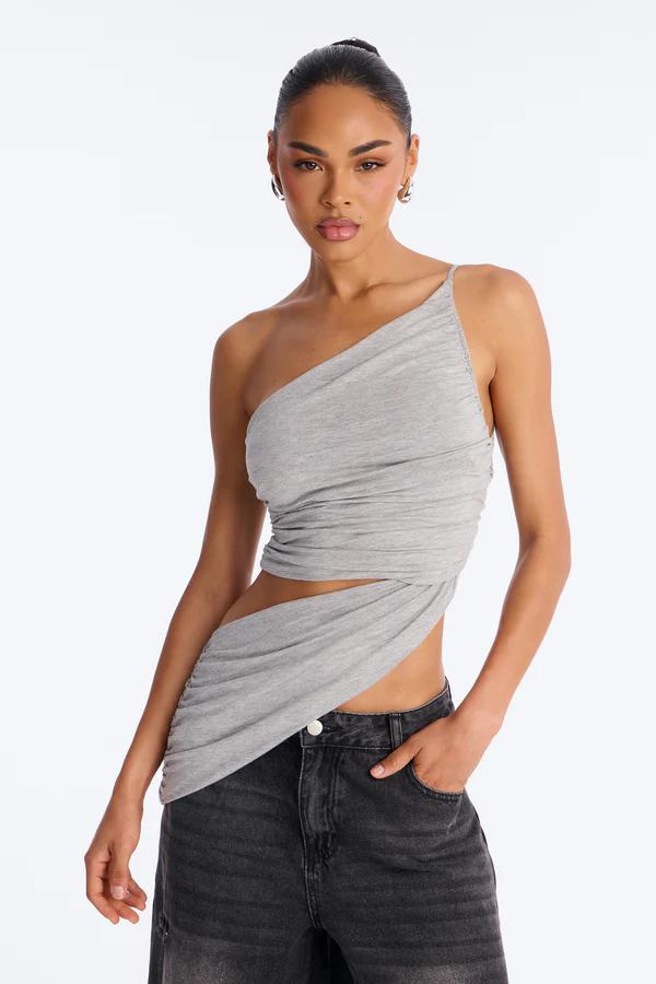 Model posing in a stylish grey one-shoulder ruched top with a midriff cutout, paired with dark wash jeans.