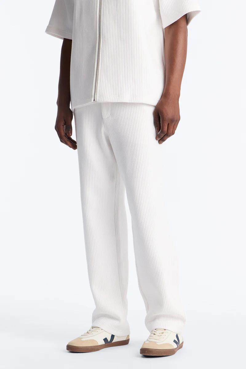 Model in off-white ribbed knit pants, a matching zippered top, and beige sneakers. Perfect for a relaxed, modern casual look.