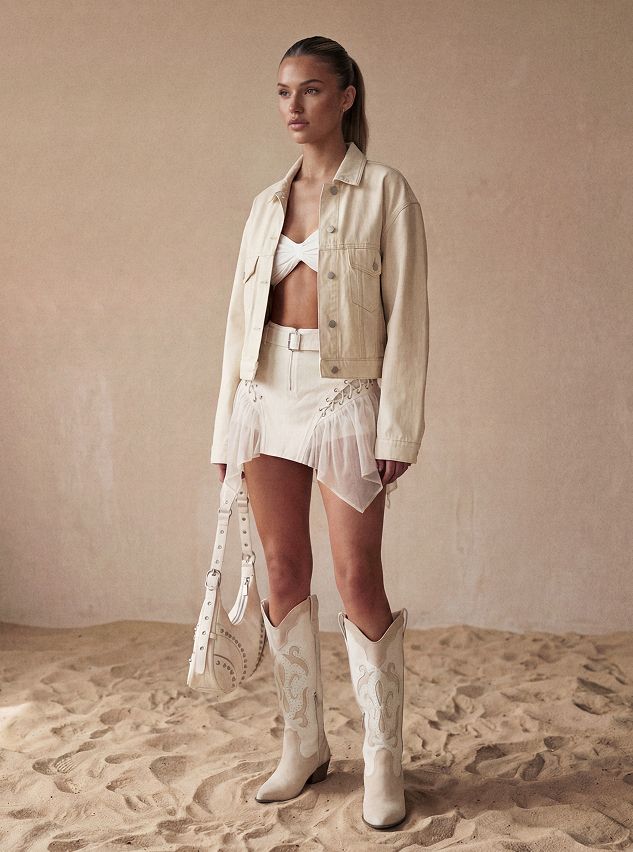 Mujer modelando chaqueta vaquera crema, top blanco, minifalda con cordones y botas vaqueras adornadas en arena. Compra nuestra nueva colección chic Western.