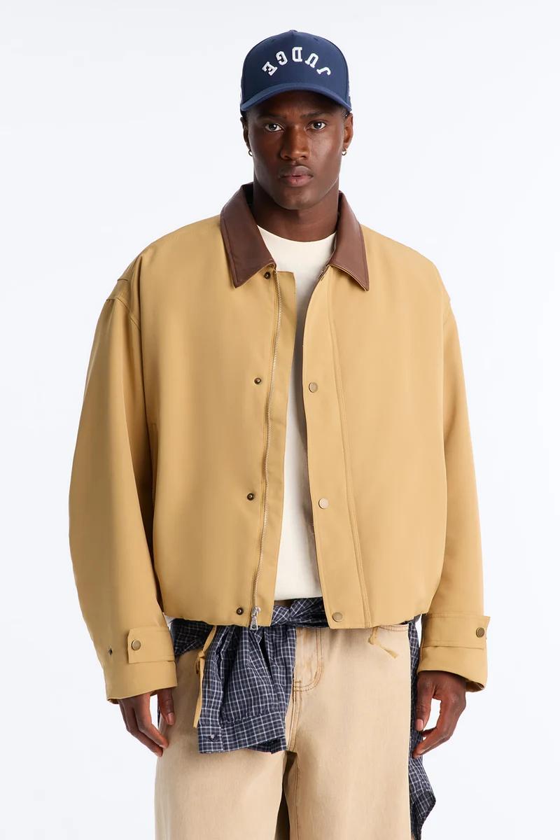 Man models a stylish tan casual jacket, brown collar, beige pants, and navy "JUDGE" cap, promoting men's fashion collection.