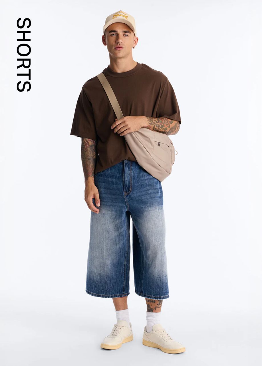 Stylish man in a brown oversized tee, faded blue wide-leg denim shorts, and beige accessories, promoting men's shorts collection.