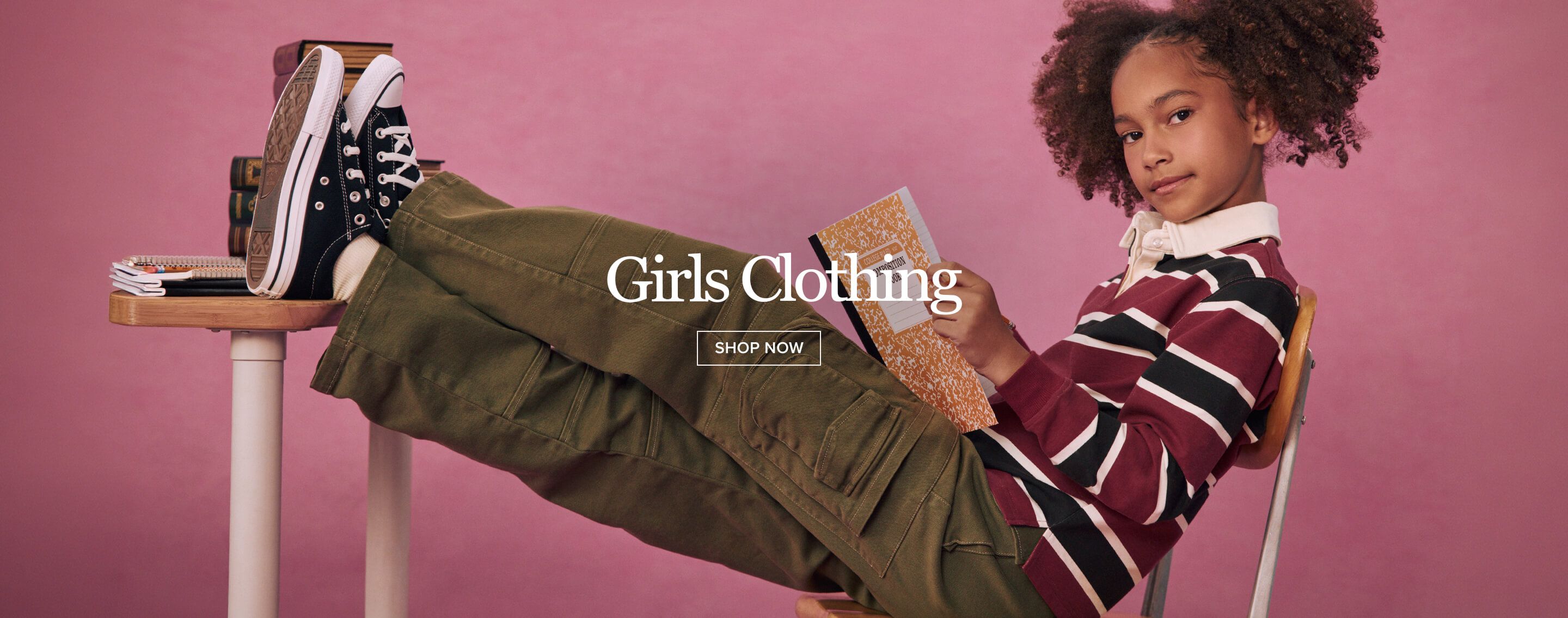 Girl wearing green cargo pants and striped polo shirt sits upside down on a school desk, holding a notebook against a pink backdrop.