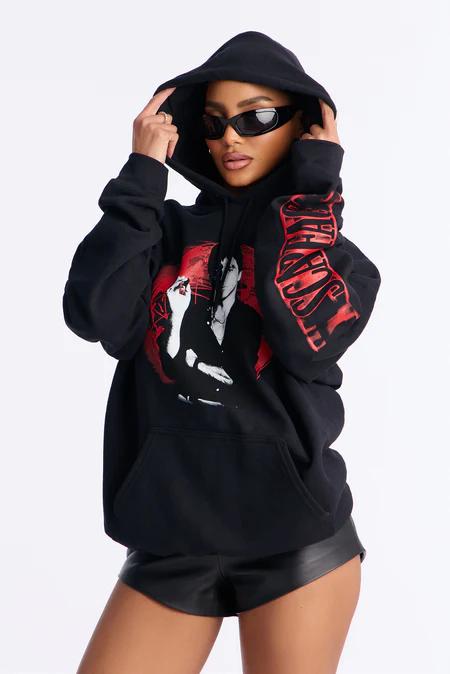 Person in a black hoodie with a red graphic featuring a figure smoking, dark sunglasses, hands adjusting the hood, black shorts.