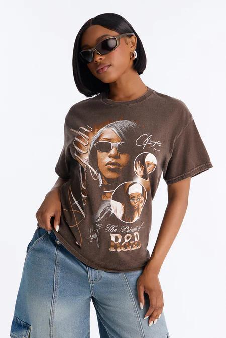 Mujer con pelo oscuro, gafas de sol y aros, viste camiseta marrón vintage de Aaliyah con retratos y texto, y vaqueros azules.