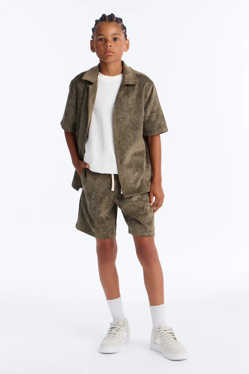 Boy in olive green paisley terry shirt & shorts set, white tee, sneakers. Casual kids' summer fashion.
