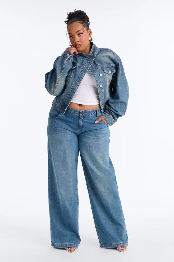 Mujer con chaqueta vaquera oversize azul claro, top corto blanco y jeans anchos, posando sobre fondo blanco.