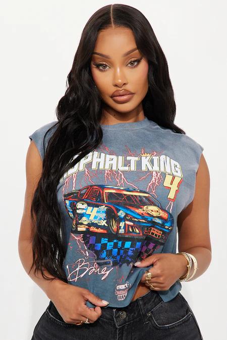 Mujer con cabello oscuro largo y ondulado, viste camiseta gris descolorida con vibrante gráfico de auto de carreras, anillos y pulseras doradas.