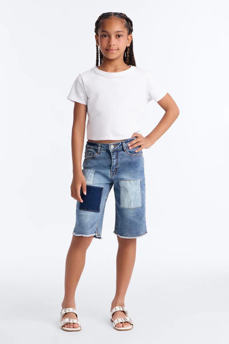 Niña con top crop blanco, bermudas de mezclilla patchwork y sandalias blancas. Moda infantil de verano.