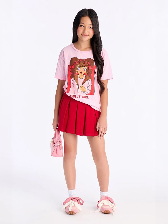 Niña sonriente modela camiseta gráfica "IT GIRL" rosa, falda plisada roja y zapatillas con lazos rosas. Colección de moda juvenil.