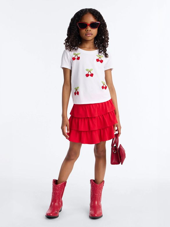 Playful girl in white cherry tee, red tiered skirt, matching red cowboy boots and sunglasses. A vibrant kids' fashion statement.