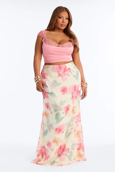 Mujer con top rosa corto de encaje y falda maxi color crema con estampado floral rosa y verde, pulseras doradas.