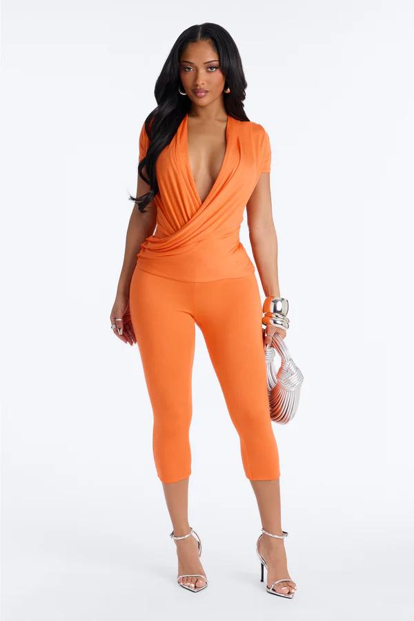 Woman showcases a trendy orange wrap plunge top and matching cropped pants set. Complemented by silver heels and an elegant bag for a chic look.