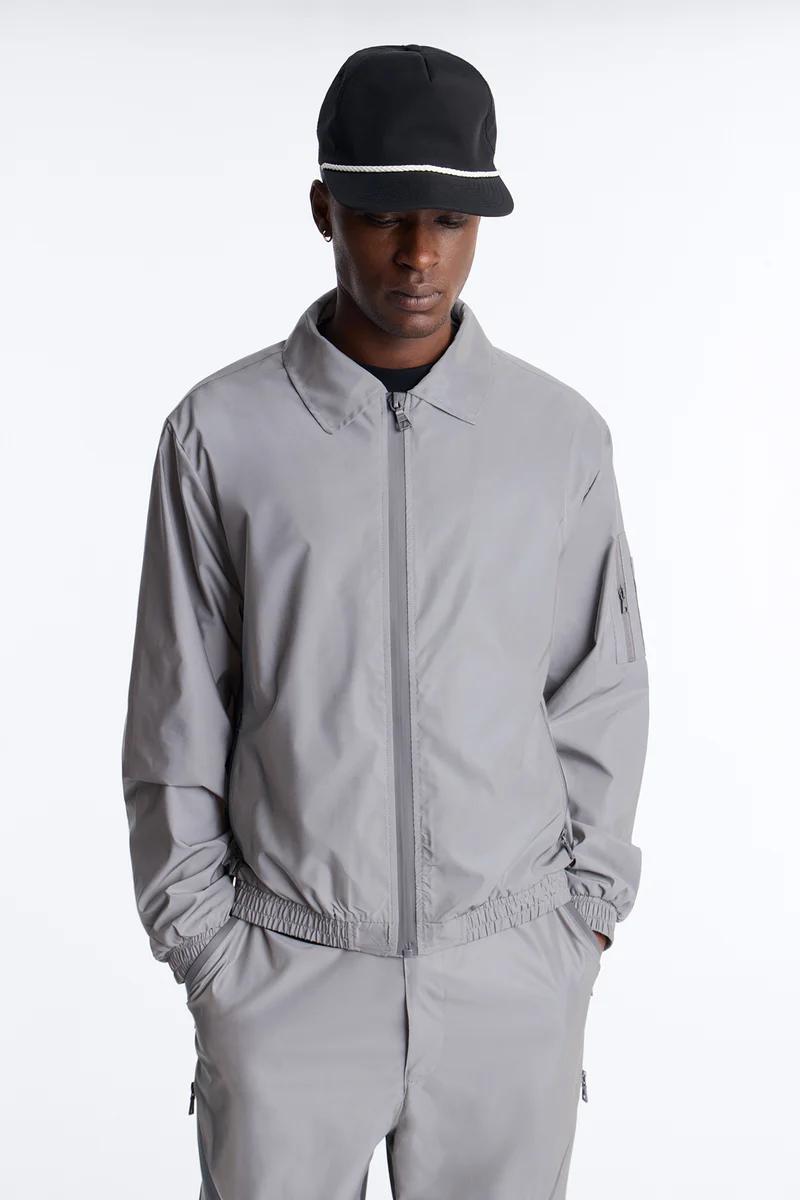 Man wears a grey full-zip tracksuit and black cap, hands in pockets, showcasing the latest men's casual sportswear.