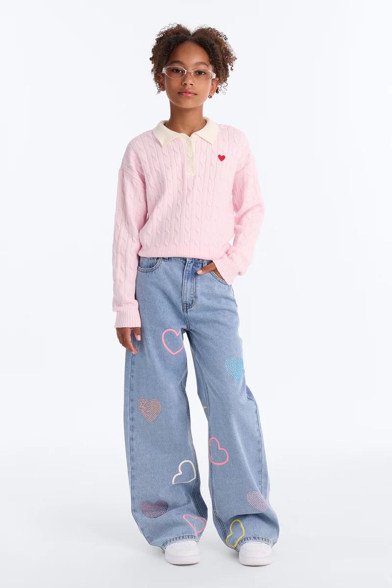 Niña con suéter rosa de punto trenzado con corazón rojo y jeans azules anchos con diseños de corazones de strass.