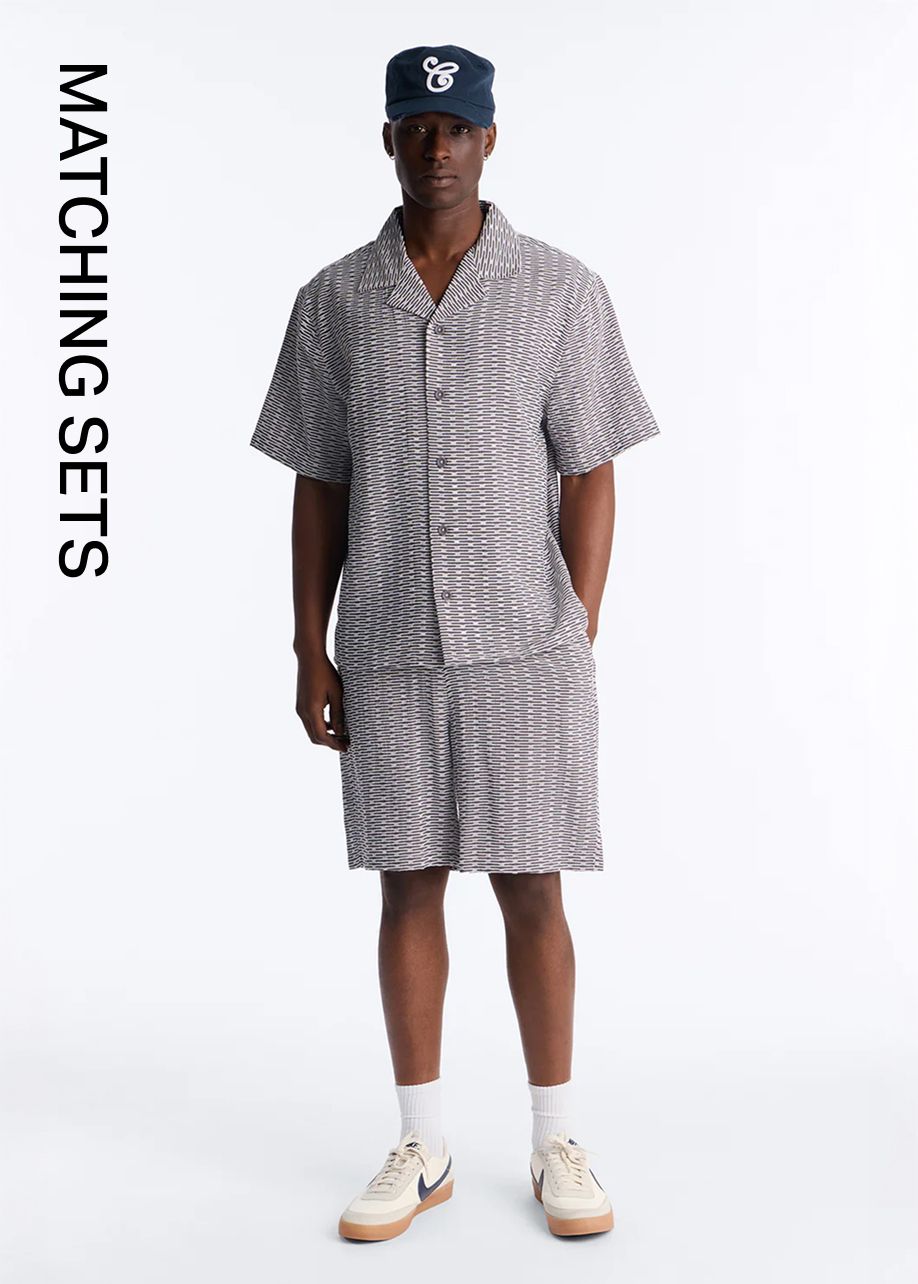 Man in a grey-white patterned matching shirt and shorts set with a navy cap. Shop coordinating men's summer outfits and matching sets.