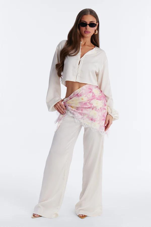 Mujer modelando camisa corta color crema, pantalones anchos y un pareo floral rosa y amarillo con encaje blanco. Lleva gafas de sol y tacones dorados.