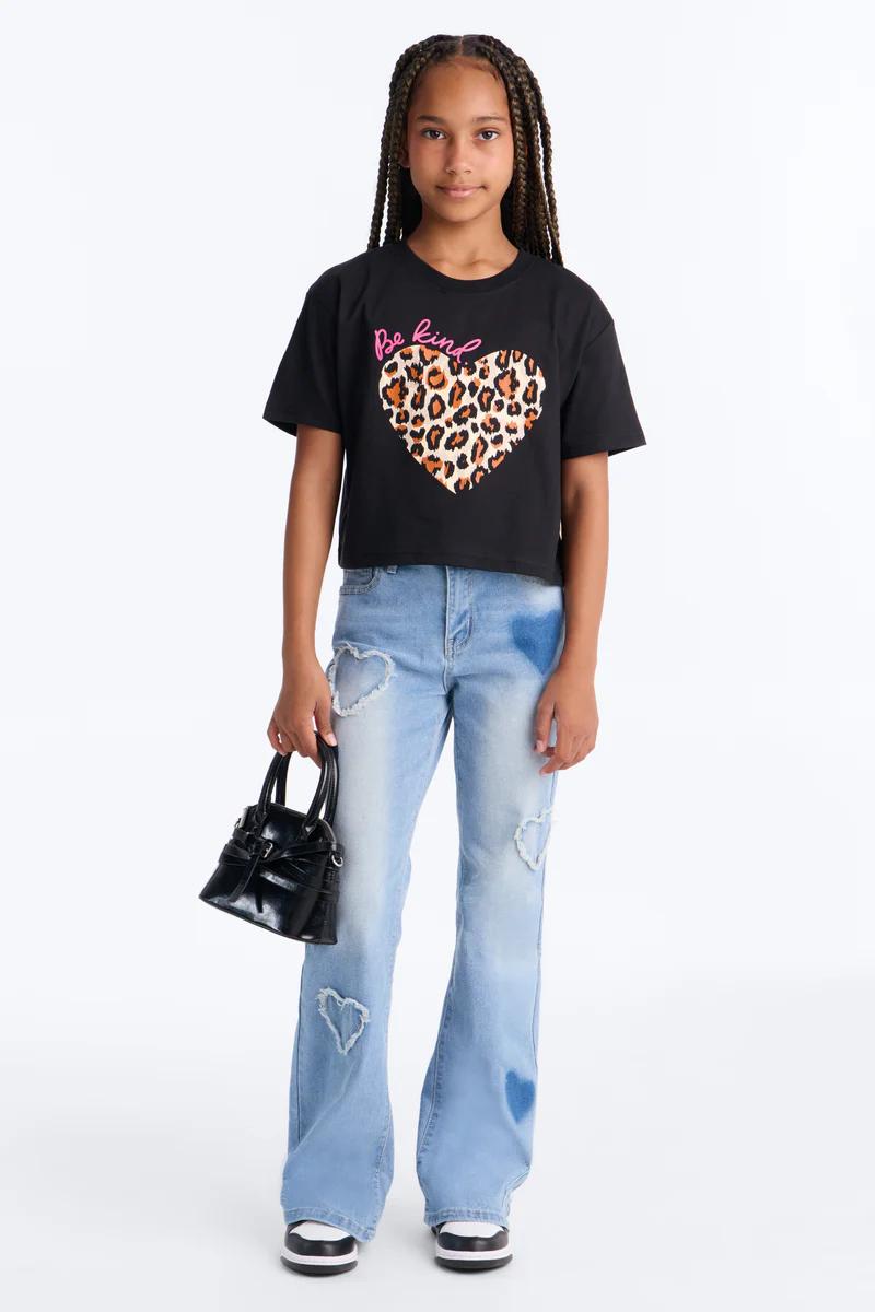 Niña a la moda con camiseta negra "Be kind" de corazón leopardo, jeans acampanados claros con corazones, mini bolso negro y zapatillas.