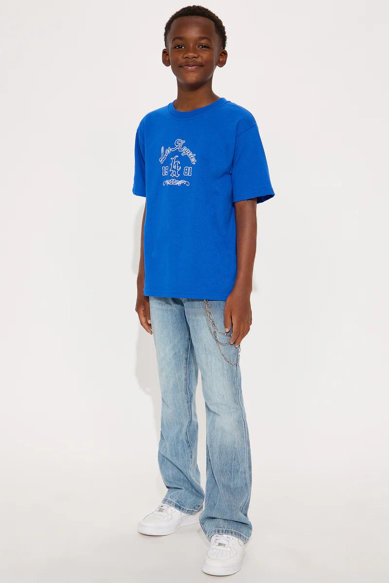 Smiling boy in blue Los Angeles 1981 graphic tee, light wash jeans with chain, & white sneakers. Kids fashion.
