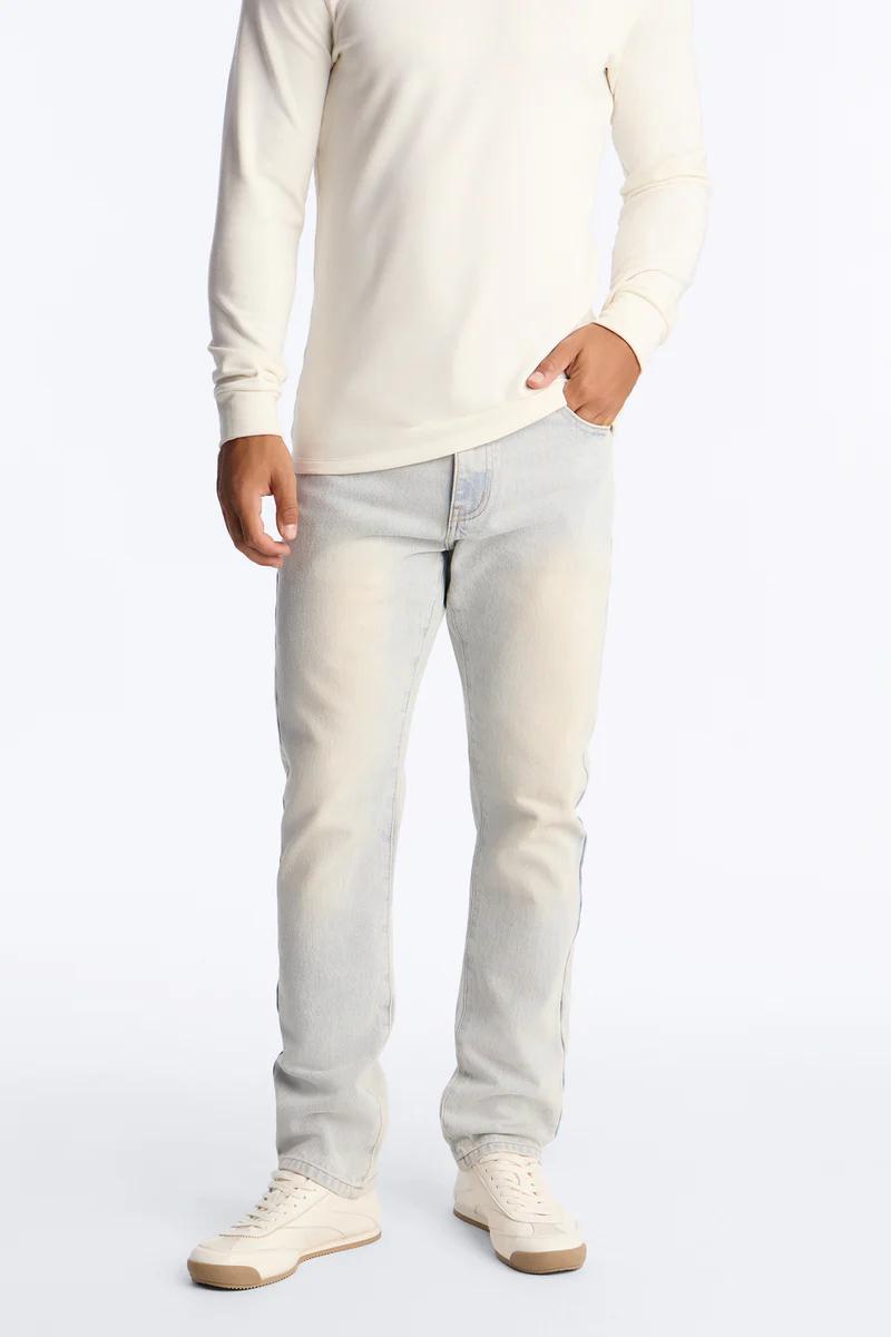 Hombre con camiseta de manga larga crema, jeans lavado claro y zapatillas. Ideal para nueva colección de moda casual masculina.