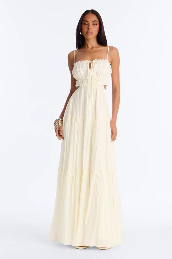 Woman in a square-neck, white maxi dress with blue floral print, bow straps, and a lace hem, holding a straw clutch.