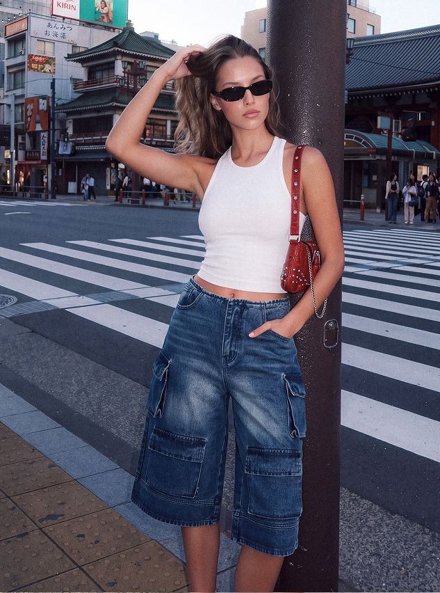 Woman models a white tank top and blue wide-leg denim cargo shorts with a red bag in an urban street scene. Highlights trendy summer fashion.