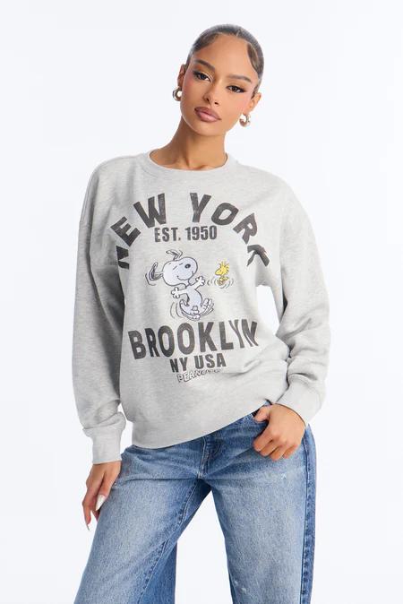 Fashionable woman posing in a light grey 'NEW YORK EST. 1950 BROOKLYN' sweatshirt featuring Snoopy & Woodstock, paired with blue jeans.