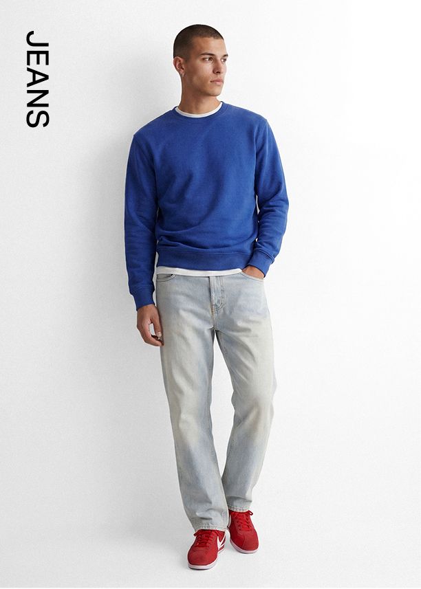 Hombre elegante con sudadera azul, jeans claros y zapatillas rojas contra pared blanca. Compra jeans para hombre de estilo casual.