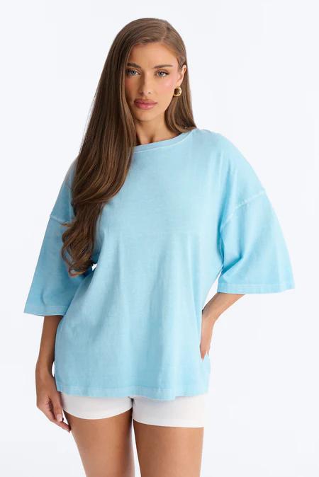 Joven con cabello castaño largo y arete dorado, vista al frente, luce camiseta azul cielo holgada de cuello redondo y shorts blancos.