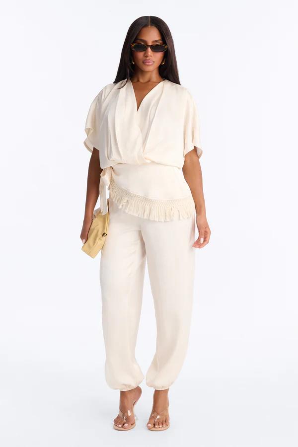 Woman in a chic cream wrap top and matching trousers with fringe detail, clear heels, and sunglasses, holding a yellow clutch. Perfect for resort wear.