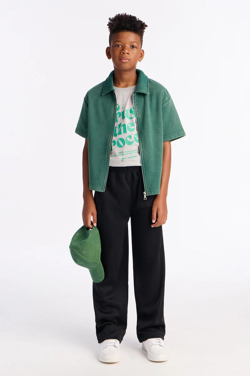 Young boy in casual green jacket, grey tee, black pants, white sneakers, holding a green cap. Kids' fashion.